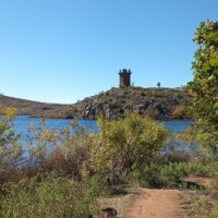 Two dogs on leashes walking a dirt path toward the Jed Johnson Watchtower Lawton Oklahoma, with the stone tower visible on a hill beside a lake under a clear blue sky.