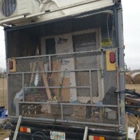 Enclosed back deck of converted bus with metal mesh gate secured shut, housing wooden boards, door, and miscellaneous items