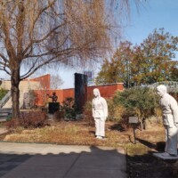 Outdoor sculpture garden near the H.U. Lee International Gate in Little Rock, featuring two white statues of hooded human figures in walking poses, a black statue in the background, and a multi-tiered fountain; surrounded by leafless trees, brick walls, and landscaped pathways under a clear blue sky.
