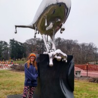 Reflection sculpture on a black pedestal in Shelby Park, Nashville—mirrored bird figure with a droplet beak, photographed beside a person in an orange hat, with trees and a construction site in the background.