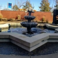 Three-tiered decorative fountain in a star-shaped basin with cascading water, set in a landscaped courtyard near the H.U. Lee International Gate in Little Rock; surrounded by brick walls, trees, and a polished black monument, creating a peaceful urban retreat.