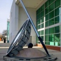 Tilted sundial sculpture leaning on a pole in front of a glass building