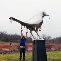 Person interacting with the Reflection sculpture in Nashville—mirrored bird structure with elongated droplets, set on a pedestal in a grassy park.