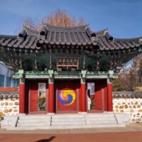 Traditional Korean-style gate at the H.U. Lee International Gate and Garden in Little Rock, Arkansas, featuring a curved tiled roof, vibrant red doors with a Sam-Taegeuk symbol, flanked by stone lion statues and patterned stone walls, with autumn trees and city buildings in the background.