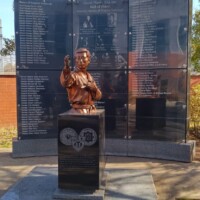 Bronze statue of a soldier in motion mounted on a black pedestal, part of the 173rd Airborne Brigade memorial near the H.U. Lee International Gate in Little Rock; backed by a granite wall engraved with names honoring veterans and fallen soldiers.