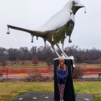 Reflection sculpture in Nashville’s Shelby Park—mirrored bird with dripping wing details on a pedestal, with a visitor in a blue jacket and patterned pants standing in front, surrounded by grass, trees, and a colorful fence.