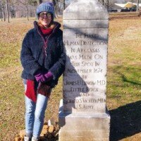 A woman in a blue knit hat and navy fleece stands beside a stone monument reading "The First Human Dissection in Arkansas Was Made On This Spot In November 1874."