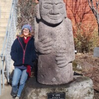 Stone Dolhareubang statue from Jeju Island, South Korea, displayed near the H.U. Lee International Gate in Little Rock; features a rounded head and folded hands, with a Korean-language plaque on the base. A bundled-up visitor stands beside it, with leafless trees and a brick building in the background.