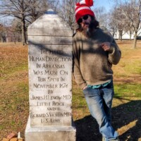Person in red-and-white striped winter hat pointing at the Human Dissection Monument in MacArthur Park — granite obelisk inscribed with names of James H. Dorman, M.D. and Ignacio S. Alvarado, M.D., marking Arkansas’s first legal dissection in 1872.