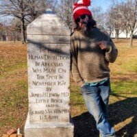 Person in red-and-white striped winter hat pointing at the Human Dissection Monument in MacArthur Park — granite obelisk inscribed with names of James H. Dorman, M.D. and Ignacio S. Alvarado, M.D., marking Arkansas’s first legal dissection in 1872.
