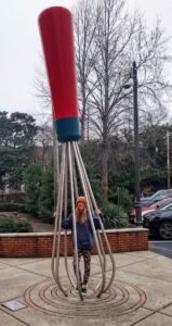 Giant red and blue whisk sculpture with a person standing beneath it in a plaza