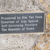 Metal plaque mounted on a rough stone surface near the H.U. Lee International Gate in Little Rock, inscribed with a dedication from Kim Tae Hwan, Governor of Jeju Special Self-Governing Province, Republic of Korea — symbolizing international friendship and cultural exchange.
