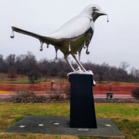 Mirrored bird sculpture titled Reflection in Nashville’s Shelby Park—appears to melt from the pedestal with metallic drips, set in a grassy area with red fencing and trees in the background