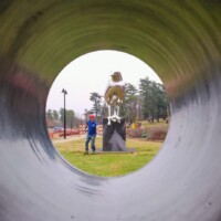 View through a circular metal tunnel framing the Reflection sculpture and a visitor in Nashville’s Shelby Park—mirrored bird statue with a droplet beak, beside a person in a blue jacket and red hat.