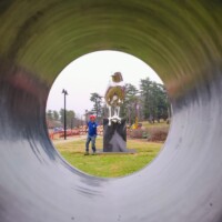 View through a circular metal tunnel framing the Reflection sculpture and a visitor in Nashville’s Shelby Park—mirrored bird statue with a droplet beak, beside a person in a blue jacket and red hat.