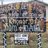 Weathered memorial sign at Billy Tripp’s Mindfield Brownsville TN, honoring Cletie with “In Memory” text, mounted on patterned poles in front of a small fenced area and building.