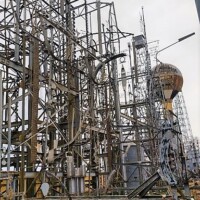 Dense arrangement of metal towers, antennas, satellite dishes, and communication equipment, supported by ladders and platforms within a fenced grassy area under cloudy skies.