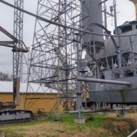 Large gray naval ship structure mounted on a platform with surrounding metal scaffolding, displayed alongside a tracked military vehicle in an outdoor exhibit area.