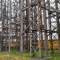 Large steel framework with vertical, horizontal, and diagonal beams forming a grid pattern, set on grassy ground with a yellow barrier in the background under cloudy skies.