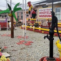 Colorful fenced display outside the Mindfield Museum with an artificial palm tree, rooster sculpture, black water‑pump statue, benches, and yellow flowers on gravel.