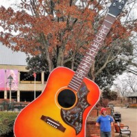 Tall orange and red Grand Ole Opry guitar sculpture with swirling designs and yellow burst accents. A person wearing a red hat and blue shirt stands beside it, with trees in autumn foliage and a balcony visible in the background.