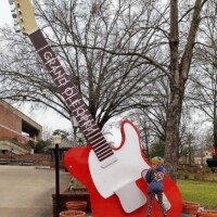 Large red and white Grand Ole Opry guitar sculpture outdoors, with flower pots around the base. A woman stands in front of it facing away, framed by trees and a nearby building.