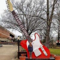 Large red and white electric guitar sculpture labeled “Grand Ole Opry,” with a person standing in front, trees and building in the background.