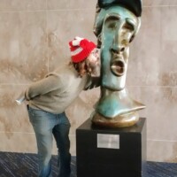 Person in a red pom-pom hat leaning in to kiss a large abstract head sculpture with exaggerated features, displayed on a black pedestal at the Knoxville Convention Center.