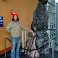 Man in a red pom-pom hat posing beside a large metal bear statue on a rocky pedestal, displayed inside the Knoxville Convention Center.