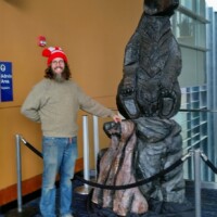 Man in a red pom-pom hat posing beside a large metal bear statue on a rocky pedestal, displayed inside the Knoxville Convention Center.
