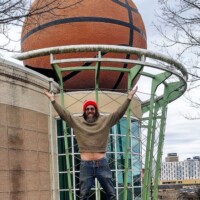 A person stands in front of the World's Largest Basketball sculpture in Knoxville, Tennessee, raising their arms to mimic the hoop structure above. The oversized fiberglass basketball is integrated into the building's architecture, appearing as if it is going through a metal hoop. Trees and a cityscape with buildings are visible in the background.