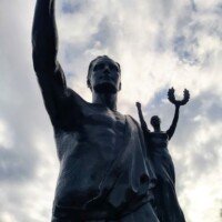 Low-angle view of a dark bronze statue of a robed figure with a raised arm against a cloudy sky