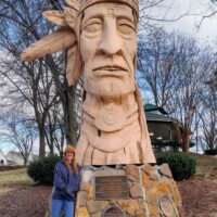 Visitor Nancy poses beside Peter Wolf Toth's 'Junaluska' Whispering Giant sculpture in Johnson City, Tennessee, showing the impressive scale of the carved wooden Native American tribute on its commemorative stone base