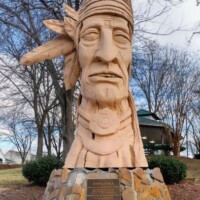 Peter Wolf Toth's 'Junaluska' Whispering Giant sculpture in Johnson City, Tennessee - a carved wooden Native American figure with traditional headband and detailed facial features, mounted on a stone base with commemorative plaques featured on a page about the Trail of the Whispering Giants