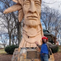 Visitor Zach stands beside Peter Wolf Toth's 'Junaluska' Whispering Giant sculpture in Johnson City, Tennessee, demonstrating the monumental scale of the carved wooden Native American tribute and its stone memorial base