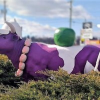 Purple and white cow statue wearing a white cap and a necklace made of miniature burgers, standing among green bushes next to a large green apple sculpture near the Purple Cow in Johnson City, TN.