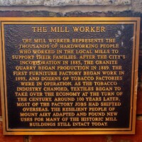 A commemorative plaque titled 'The Mill Worker,' mounted on a brick wall. The text honors the hardworking individuals who labored in local mills to support their families. It describes the evolution of Mount Airy’s economy—from granite quarrying and tobacco production in the late 1800s to the rise of textiles in the 20th century—and acknowledges the community’s resilience in repurposing historic mill buildings after factory jobs moved overseas.