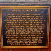 A commemorative plaque titled 'The Mill Worker,' mounted on a brick wall. The text honors the hardworking individuals who labored in local mills to support their families. It describes the evolution of Mount Airy’s economy—from granite quarrying and tobacco production in the late 1800s to the rise of textiles in the 20th century—and acknowledges the community’s resilience in repurposing historic mill buildings after factory jobs moved overseas.