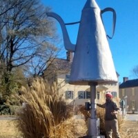 Oversized silver teapot sculpture known as The Mickey Coffee Pot, standing tall in Winston-Salem with a person in a red hat nearby for scale
