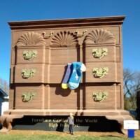 The World's Largest Chest of Drawers, a towering 38-foot furniture landmark in High Point, North Carolina. Its ornate gold handles and playful oversized socks hanging from an open drawer highlight the city's rich furniture heritage. The bold lettering reads 'Furniture Capital of the World,' reinforcing its iconic status. A visitor stands in front, showing its impressive scale.