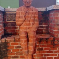 A life-sized brick sculpture of a seated man integrated into the wall, dressed in a suit and tie, with his right hand resting as if holding a small object. A commemorative plaque mounted above the sculpture provides historical context, though the text is not fully visible in this image.