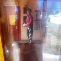 Interior view of The Last Shell-Shaped Shell Service Station, seen through aged glass with visible distortions from sun exposure, capturing vintage framed pictures and a door with a yellow sign.