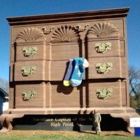 The World's Largest Chest of Drawers in High Point, North Carolina—a towering, 38-foot dresser with ornate gold handles and two oversized socks hanging from an open drawer. The landmark celebrates the city's furniture heritage with bold lettering that reads 'Furniture Capital of the World.' A visitor stands nearby, highlighting its incredible scale.