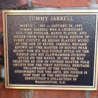 A commemorative plaque honoring Tommy Jarrell, the legendary old-time fiddler and banjo player from the Round Peak region of Surry County. The plaque details his early start in music, his distinctive fiddle and banjo styles, and his recognition as a National Heritage Fellow in 1982. It also notes that his fiddle is now part of the Smithsonian Institution’s collection.
