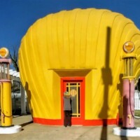 The Last Shell-Shaped Shell Service Station, shaped like a bright yellow seashell with a red door, flanked by vintage Shell-branded gas pumps in pastel yellow and pink.