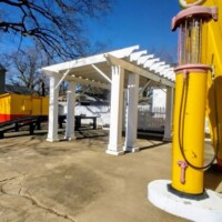 A vintage yellow gas pump stands in front of The Last Shell-Shaped Shell Service Station, with a white pergola structure and black ramps behind it. A green house and a yellow shed sit further back, framed by bare trees and a cracked concrete surface under a bright blue sky.