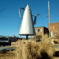Oversized silver Mickey Coffee Pot sculpture in Winston-Salem, sitting atop a pedestal surrounded by tall, dry grass with brick buildings and power lines in the background."