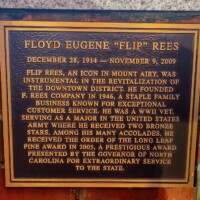 A commemorative plaque honoring Floyd Eugene 'Flip' Rees, mounted on a brick wall. The plaque details his contributions to Mount Airy’s downtown revitalization, his founding of F. Rees Company in 1946, his decorated WWII service as a major in the U.S. Army with two Bronze Stars, and his receipt of the Order of the Long Leaf Pine award in 2005 for extraordinary service to North Carolina.
