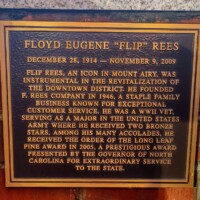 A commemorative plaque honoring Floyd Eugene 'Flip' Rees, mounted on a brick wall. The plaque details his contributions to Mount Airy’s downtown revitalization, his founding of F. Rees Company in 1946, his decorated WWII service as a major in the U.S. Army with two Bronze Stars, and his receipt of the Order of the Long Leaf Pine award in 2005 for extraordinary service to North Carolina.