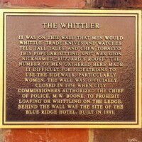 A mounted plaque titled 'The Whittler,' describing the historical significance of a local gathering spot known as Buzzard’s Roost. The plaque details how men used to whittle, trade knives and watches, tell tall tales, and chew tobacco on this wall, leading to sidewalk congestion before its official closure in 1956